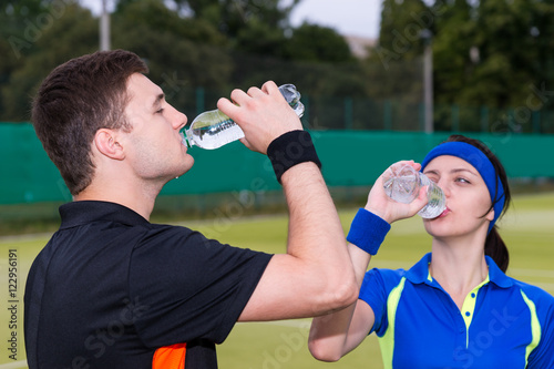 Athletic couple of tennis players drinking water after match out