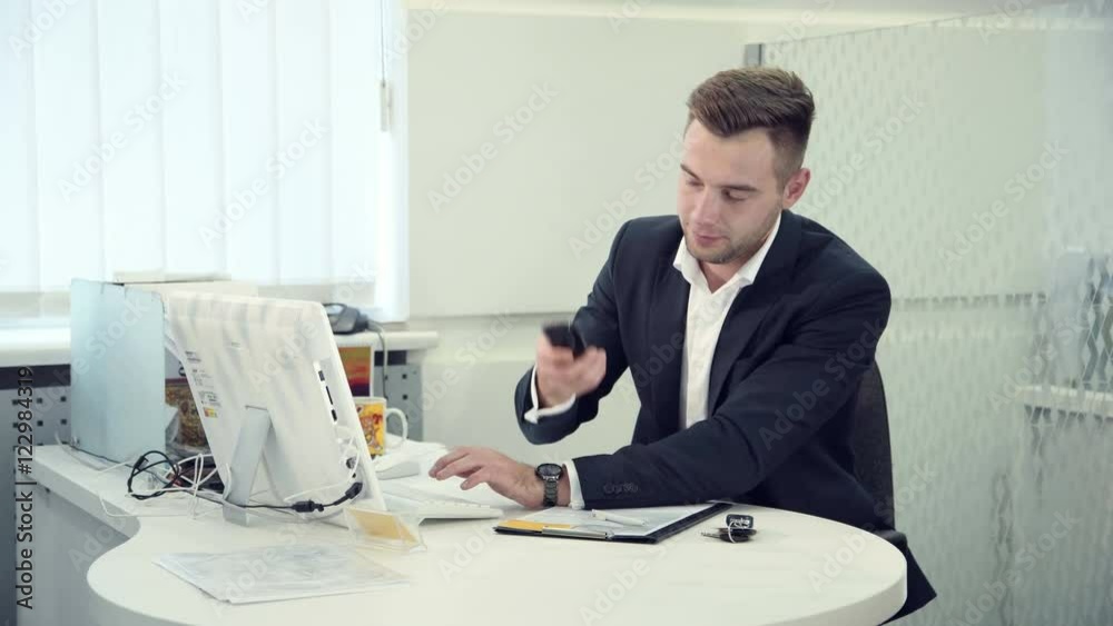 Hardworking businessman typing on a computer as he sits at a table in a modern white office, side view, then he recieved a good news by the phone. Hardworking businessman typing on a computer as he sits at a table in a modern white office, side view, then he recieved a good news by the phone.