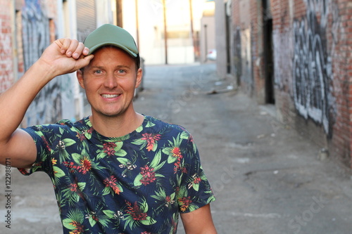 Caucasian American Man Casual Street Fashion in New York, wearing blue flower patterned shirt, green cap, standing by graffiti wall, smiling.