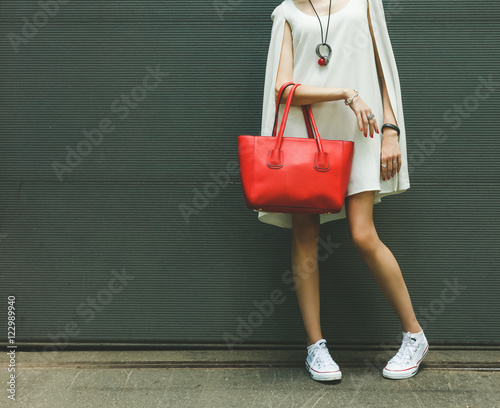 Fashionable beautiful big red handbag on the arm of the girl in a fashionable white dress, posing near the wall on a warm summer night. Warm color.