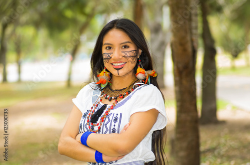 Beautiful Amazonian woman with indigenous facial paint and white traditional dress posing happily for camera in park environment, forest background photo