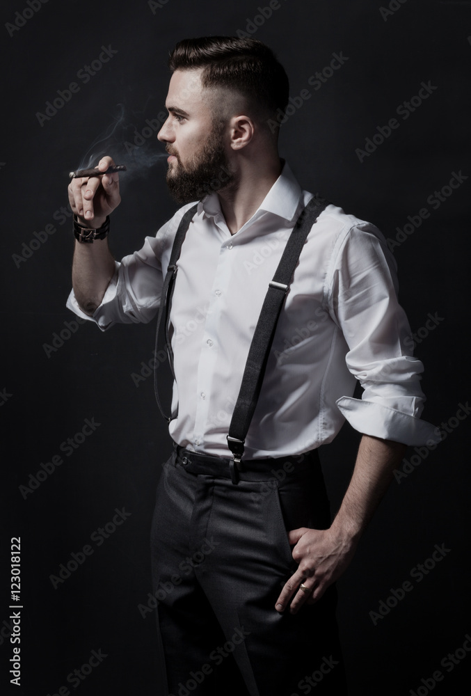 Φωτογραφία Young stylish bearded man in a white shirt and suspenders smoking a cigar in a dark room