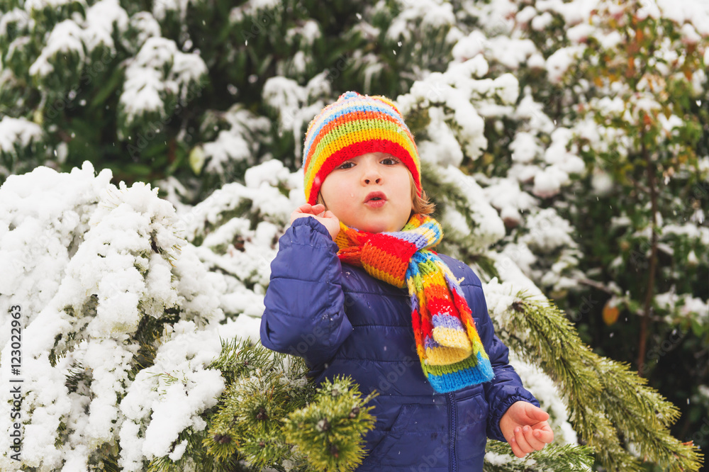 Outdoor portrait young 4 year old boy wearing blue jacket, colourful set of handmade knitted hat and scarf, enjoying winter time
