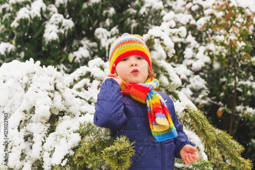 Outdoor portrait young 4 year old boy wearing blue jacket, colourful set of handmade knitted hat and scarf, enjoying winter time