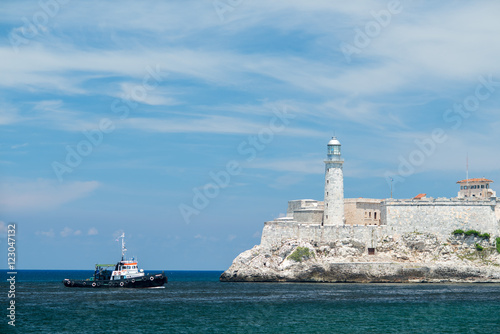 Small ship is floating in the Atlantic Ocean. Lighthouse on a fortress is near an entrance to Havana bay at summer sunny day.
