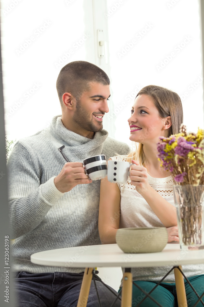 Happy young couple is sitting at the dining table, drinking coffee and looking at each other.