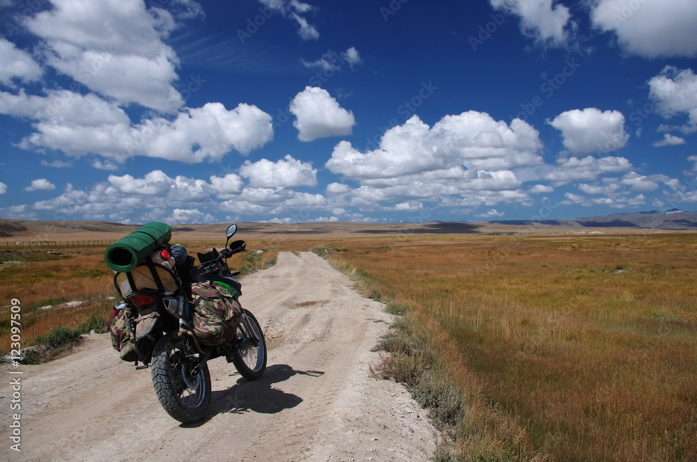 Enduro motorcycle traveler with suitcases standing on a dirt road vanishing into the skyline under a blue sky with white clouds, Plateau Ukok, Altai mountains, Siberia, Russia. Enduro motorcycle traveler with suitcases standing on a dirt road vanishing into the skyline under a blue sky with white clouds, Plateau Ukok, Altai mountains, Siberia, Russia.