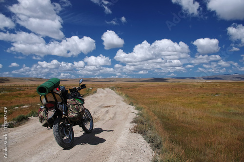 Enduro motorcycle traveler with suitcases standing on a dirt road vanishing into the skyline under a blue sky with white clouds  Plateau Ukok  Altai mountains  Siberia  Russia.