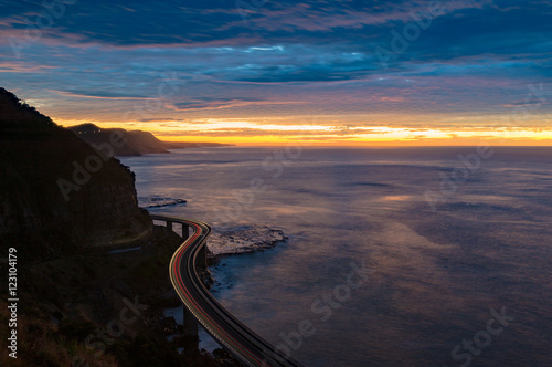 Sea Cliff Bridge on sunrise with moving traffic and dramatic beautiful sky and ocean shore on the background. The Bridge is part of NSW Grand Pacific scenic route