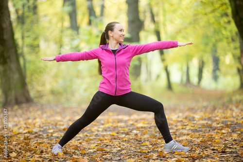 Sporty beautiful smiling young woman practicing yoga, standing in Warrior II posture, Virabhadrasana Two pose, working out outdoors on autumn day wearing sportswear sweatshirt. Full length, side view