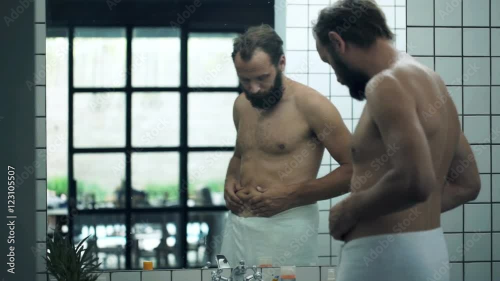 Unhappy, sad man checking his belly in front of mirror in bathroom
