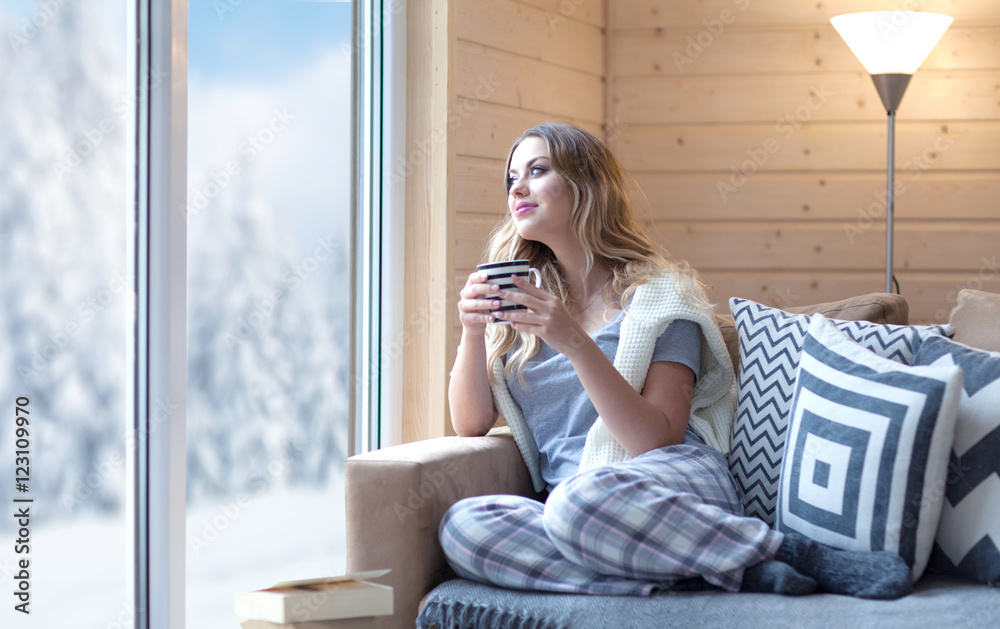 Young beautiful blonde woman with cup of coffee sitting home in living room by the window. Winter snow landscape view. Lazy day off concept - obrazy, fototapety, plakaty 