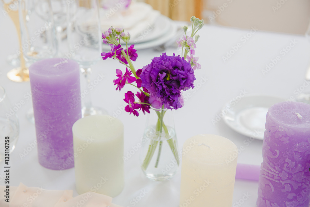 Violet and white candles surround  a glass with flowers which st