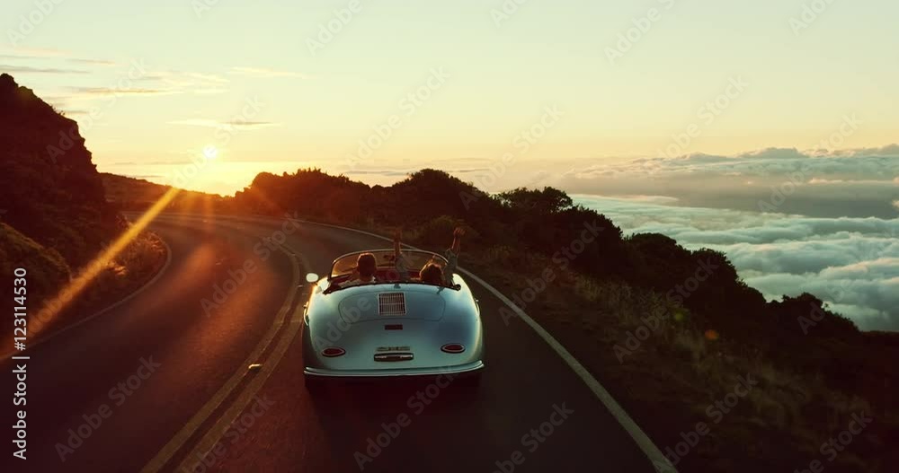 Happy couple driving on country road into the sunset in classic vintage sports car