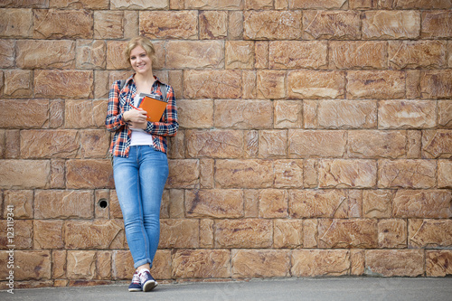 Full length portrait of a smiling student girl against the brick wall with textbooks. Back to school concept photo, looking at the camera, copy space