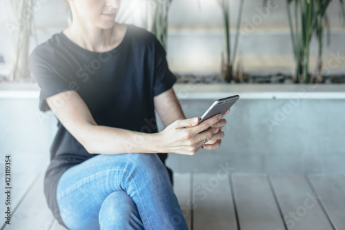 Young woman holding gray smartphone while sitting on bench with wooden texture in room.Girl uses the gadget.Girl,dressed in a black T-shirt and blue jeans sitting and surf the Internet on smartphone.