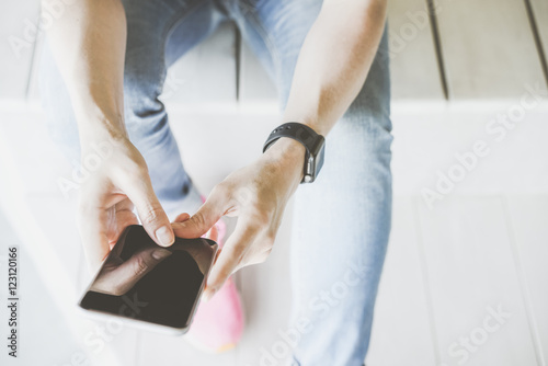 Close-up of smartphone in hands of girl sitting on bench with wooden texture.Girl dressed in blue jeans and pink shoes. Smartwatch on a female hand. A young woman using a digital gadget. Mock up.