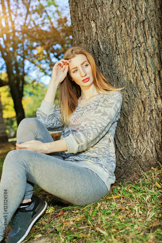 Young beautiful girl with blue eyes and blonde hair sitting near a tree