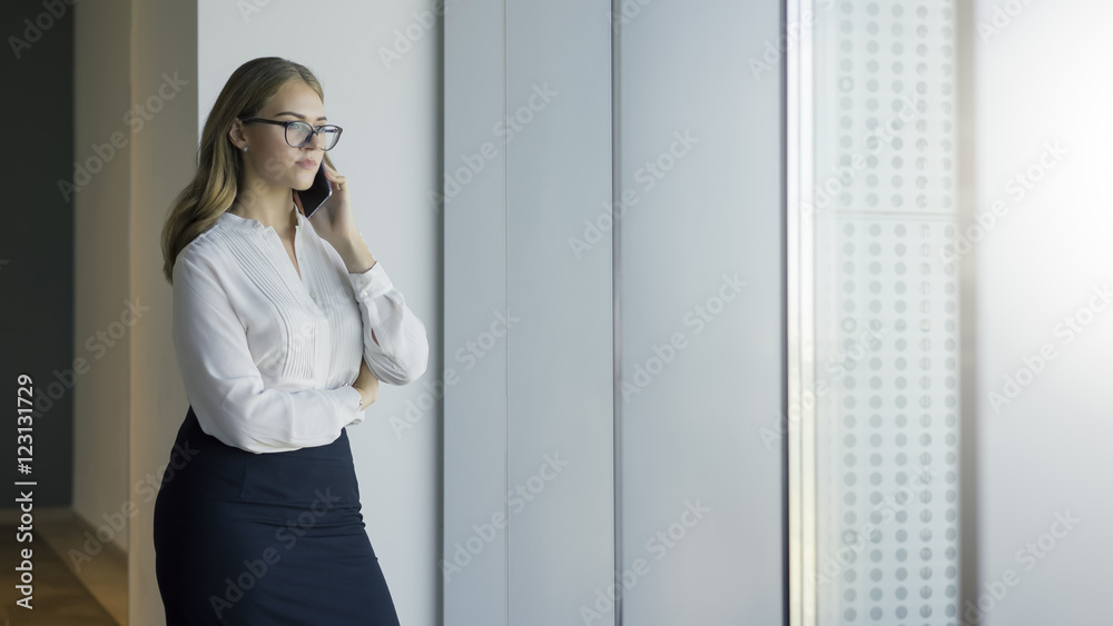A young business woman talks on the phone. Girl in a black skirt and white blouse, standing at the window and talking on a smartphone. A young business woman talks on the phone. Girl in a black skirt and white blouse, standing at the window and talking on a smartphone.