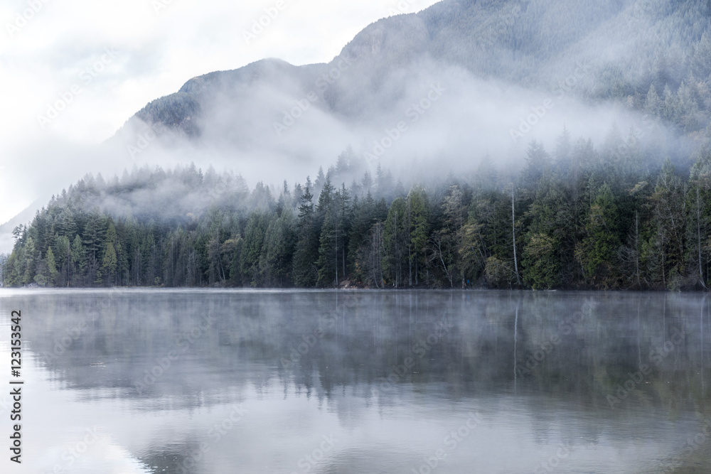 Buntzen Lake, Vancouver, British Columbia, Canada. Foggy lake in the morning. Evergreen forest reflection and mountain background.