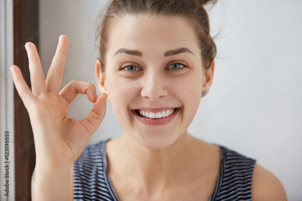 Young European brunette female showing OK-gesture with her fingers. Happy woman in striped top smiling with blue eyes. Her white-teeth mouth and happy face prove everything goes according to plan. Young European brunette female showing OK-gesture with her fingers. Happy woman in striped top smiling with blue eyes. Her white-teeth mouth and happy face prove everything goes according to plan.