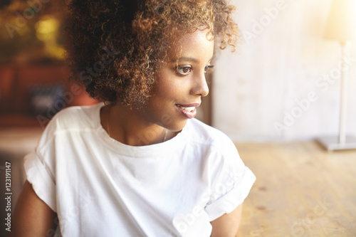 African woman in white trendy shirt waiting for friends to join her at cozy cafe. Profile portrait of young black female with pretty face and Afro hairstyle looking through window at city landscape photo