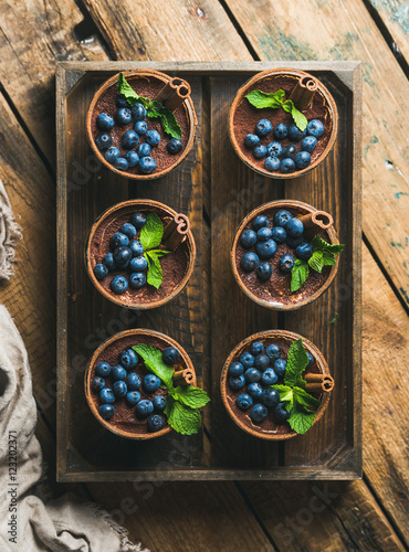 Homemade Tiramisu dessert in glasses with cinnamon sticks, mint leaves and fresh blueberries served in wooden tray over rustic wooden background, top view, vertical composition