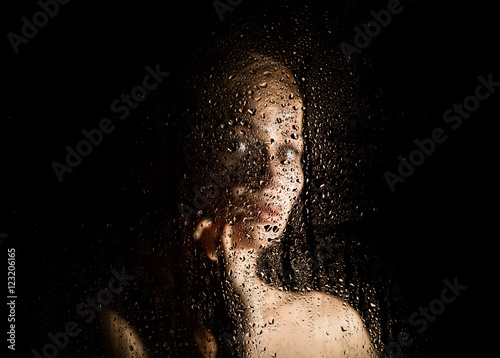 sexy young woman, posing behind transparent glass covered by water drops. melancholy and sad female portrait