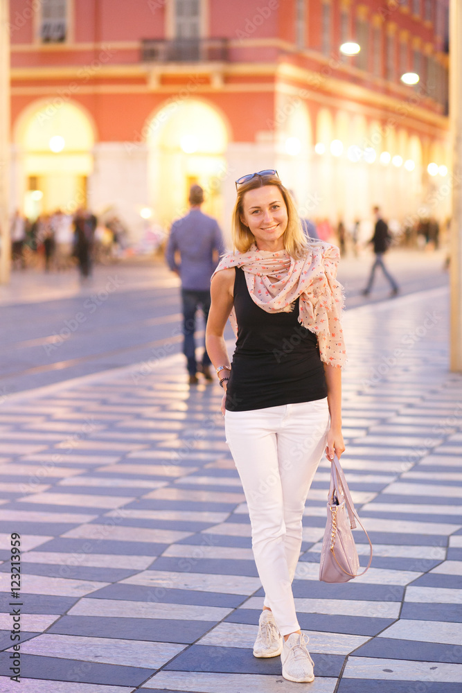 Gorgeous woman in white trousers stands on the square illuminate