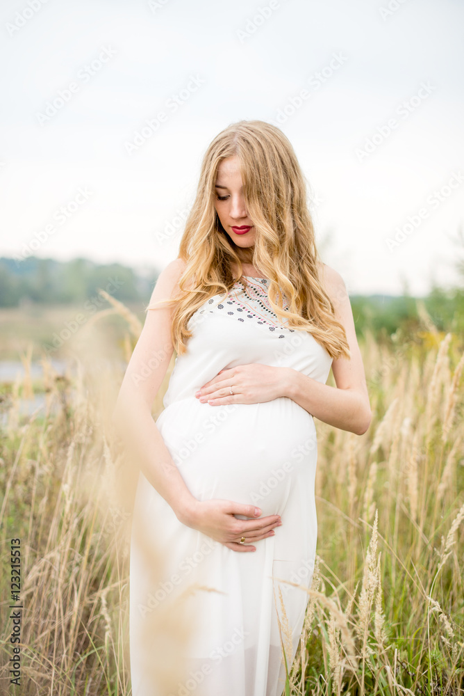 Yonge pregnant woman in a white dress with long blond hair in tall grass Yonge pregnant woman in a white dress with long blond hair in tall grass