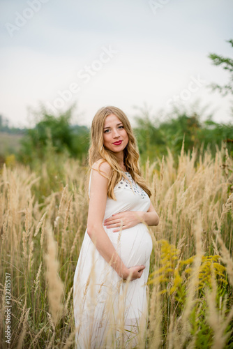 Happy and yonge pregnant woman in a white dress with long blond hair in tall grass