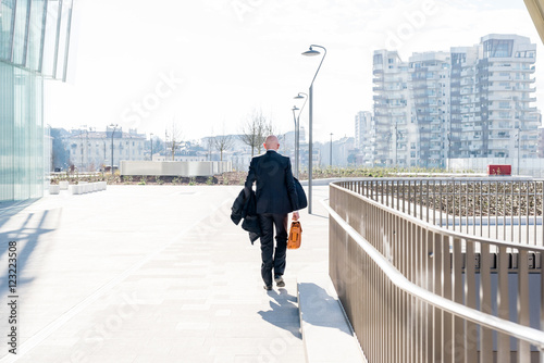 Back view of middle-age businessman holding briefcase walking outdoor in back light - business, entrepreneurship, elegant concept
