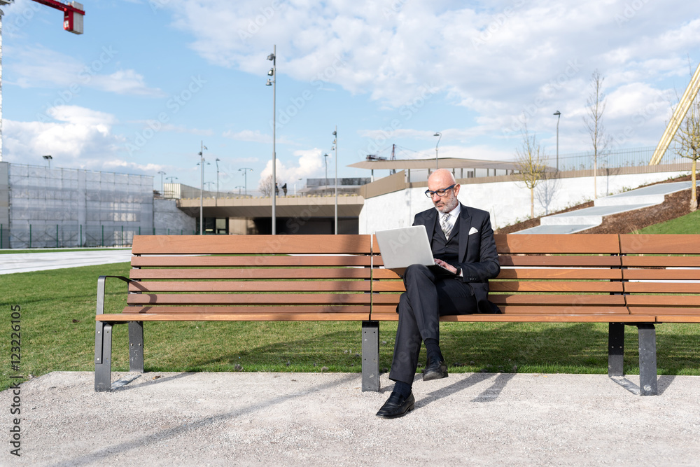 Midlle age caucasian businessman sitting on bench in the city using laptop - working, technology, business concept Midlle age caucasian businessman sitting on bench in the city using laptop - working, technology, business concept