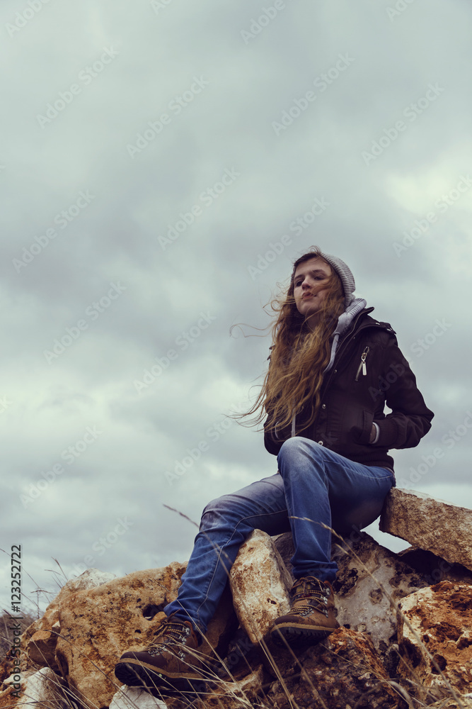 Fotografia do Stock: Mujer joven con ropa informal de abrigo sentada sobre  la cima de una montaña de rocas con el cielo nublado de fondo