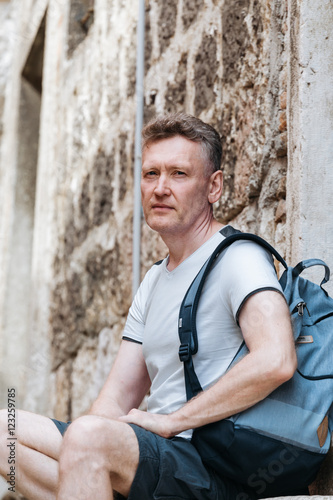 Stylish tourist. Man dressed in a white shirt and blue shorts with a blue backpack over his shoulder. Sitting on the steps of an old European city