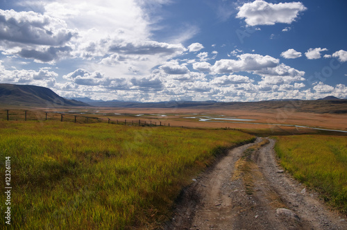 Road along the border line on a highland mountain plateau with the green grass at the background of the valley of white river under a blue sky with white clouds, Plateau Ukok, Altai, Siberia, Russia