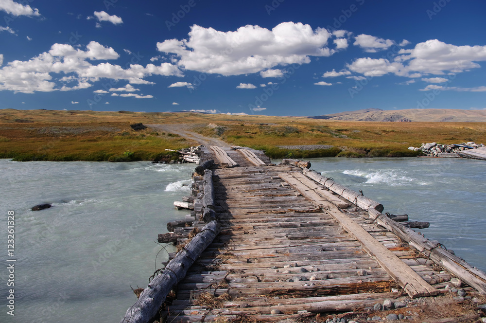 Log wooden bridge across the rapid white mountain river on a background of yellow hills under blue sky with white clouds Plateau Ukok, Altai, Siberia, Russia
