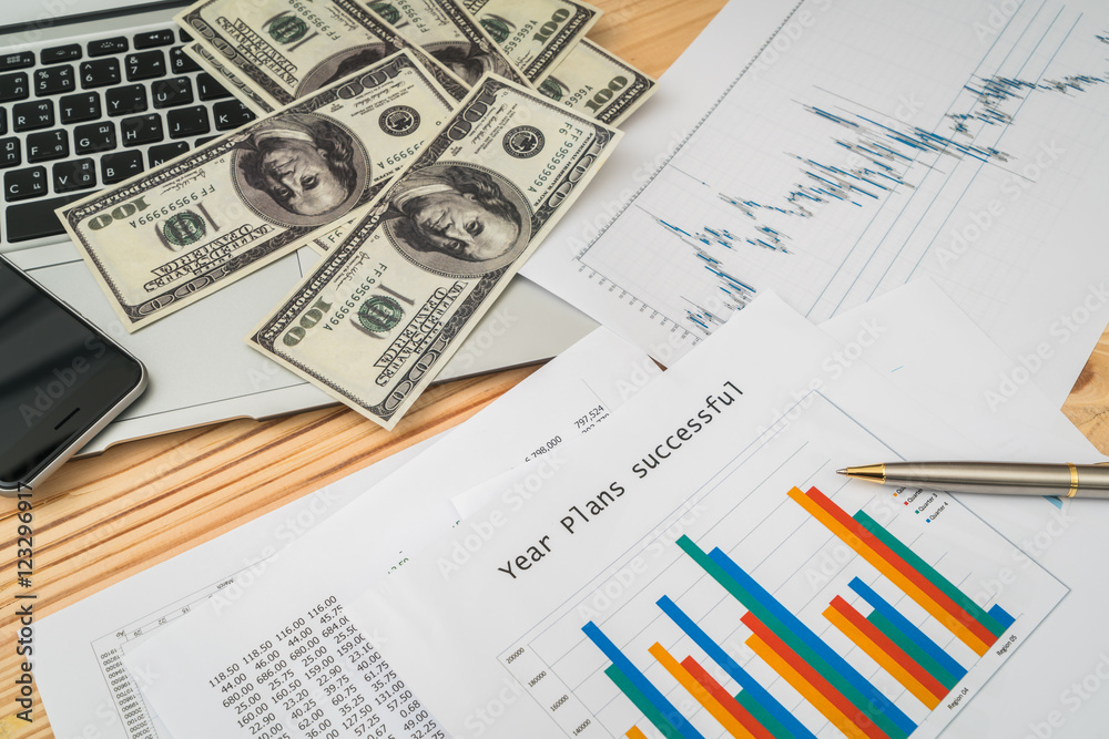 Financial charts on the table with laptop , phone ,dollars bankn Financial charts on the table with laptop , phone ,dollars bankn