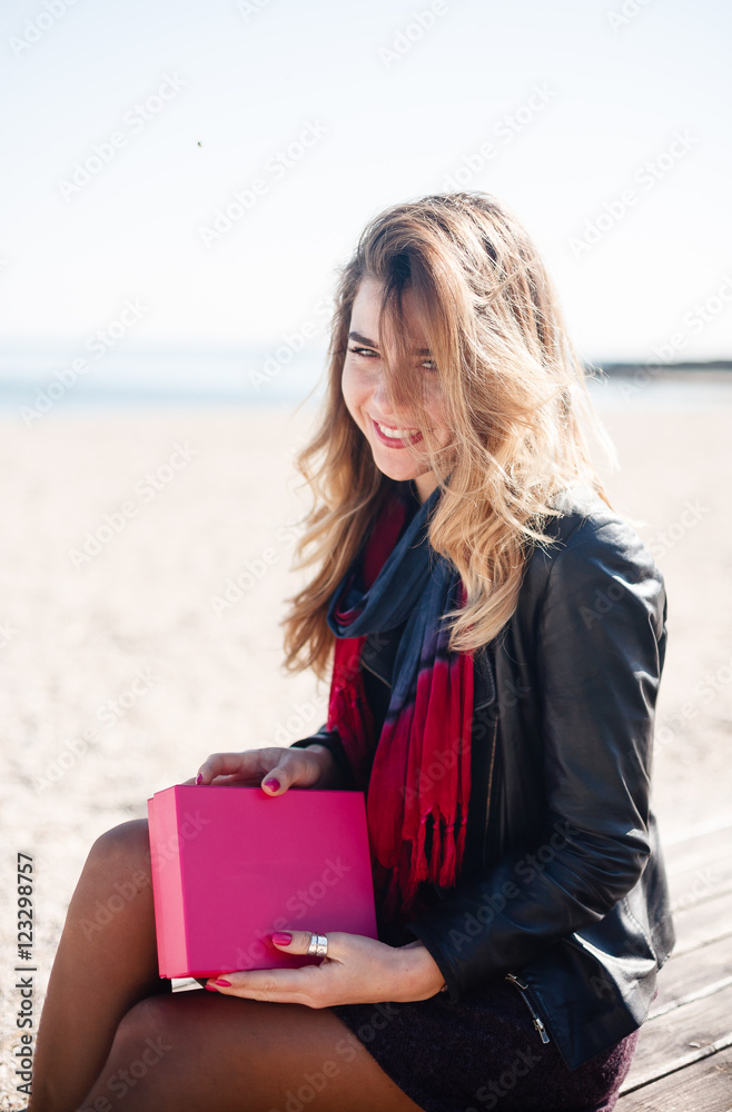 Young beautiful girl in dark clothes sitting on an empty beach and holding a pink box