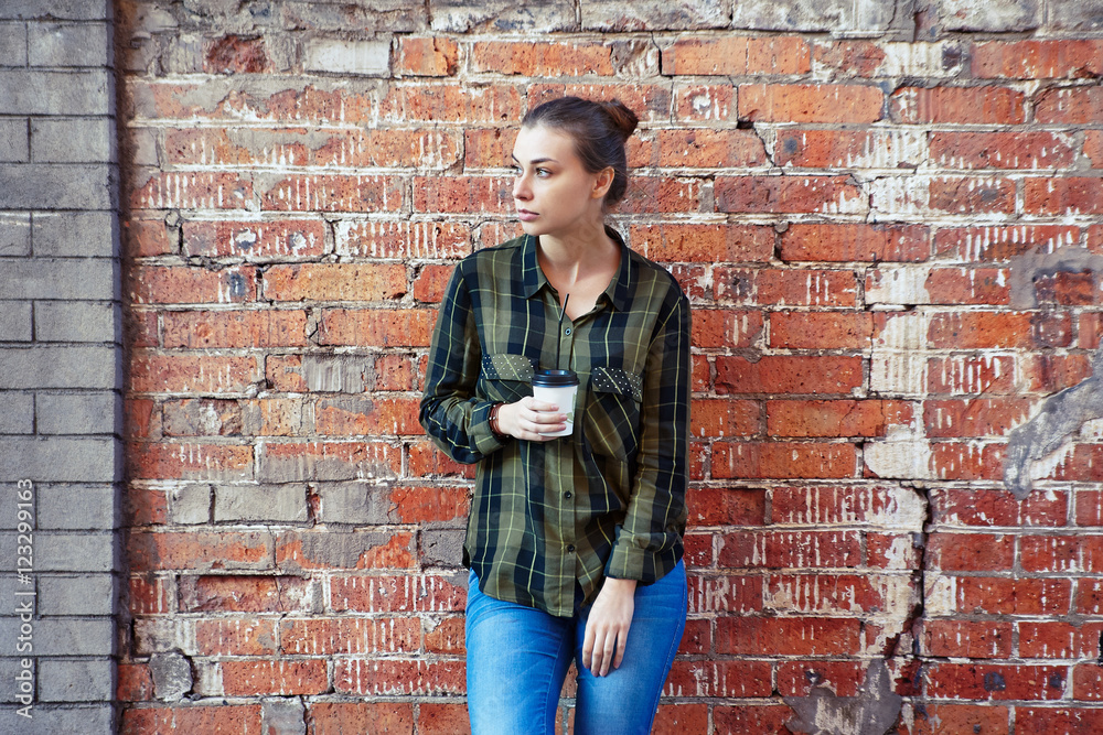 young woman in a plaid green shirt standing disposable coffee cup on the background  the red brick wall