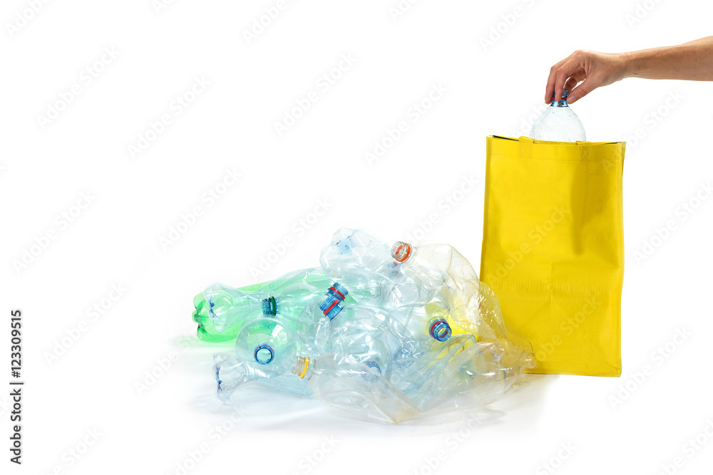 Pile of plastic bottles is lying on the white background. Woman's hand is inserting plastic bottle into an empty yellow bag. All potential trademarks are removed.