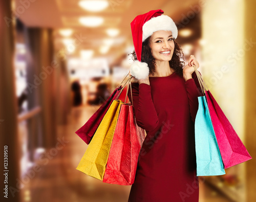 Beautiful happy woman in Santa hat with shopping bags on blurred market background. Christmas shopping concept.
