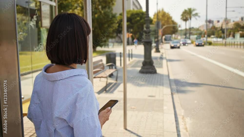 Beautiful brunette girl waits her bus on stop station in summer time and checking arrival schedule on her smartphone