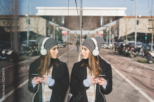 Young beautiful caucasian blonde girl listening music with headphones and smart phone hand hold, reflecting on a mirror window - music, technology, relaxing concept photo