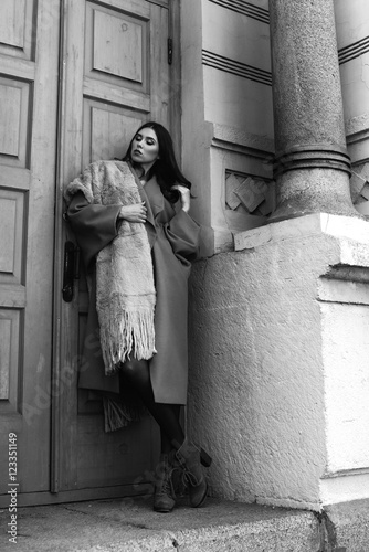 Beautiful girl with hair and make-up, walking around the city / standing near a big blue door next to the columns. Dressed in a long warm beige coat and a fur scarf. Street style.