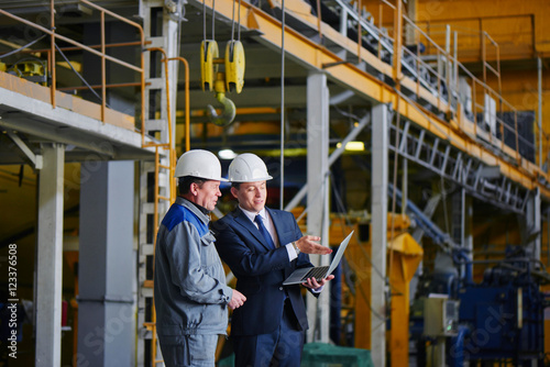 The man in the suit and the helmet holds the portable computer and shows up on the screen to the worker in overalls in an industrial building