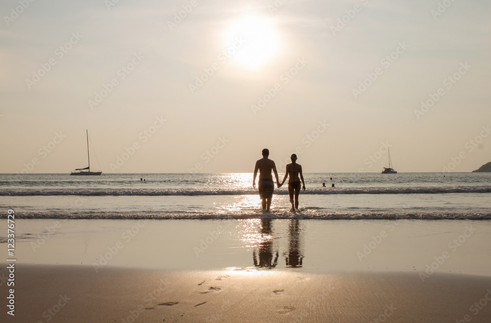 Silhouette of couple walking on beach at sunset holding hands. Horizontally framed shot.
