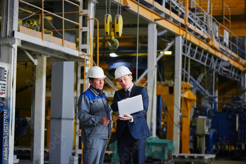 The man in the suit and the helmet holds the portable computer and shows up on the screen to the worker in overalls in an industrial building