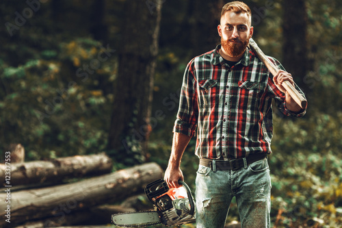 Lumberjack worker standing  in the forest with axe and chainsaw photo