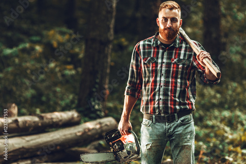 Lumberjack worker standing  in the forest with axe and chainsaw